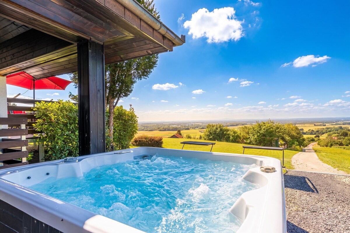 A hot tub with a wooden deck and a red umbrella, surrounded by greenery and a scenic view.