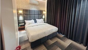 A modern hotel room with a large bed, a headboard with built-in lighting, a ceiling fan, and a dark curtain on the window. - Eminence the Corbett (Ramnagar)