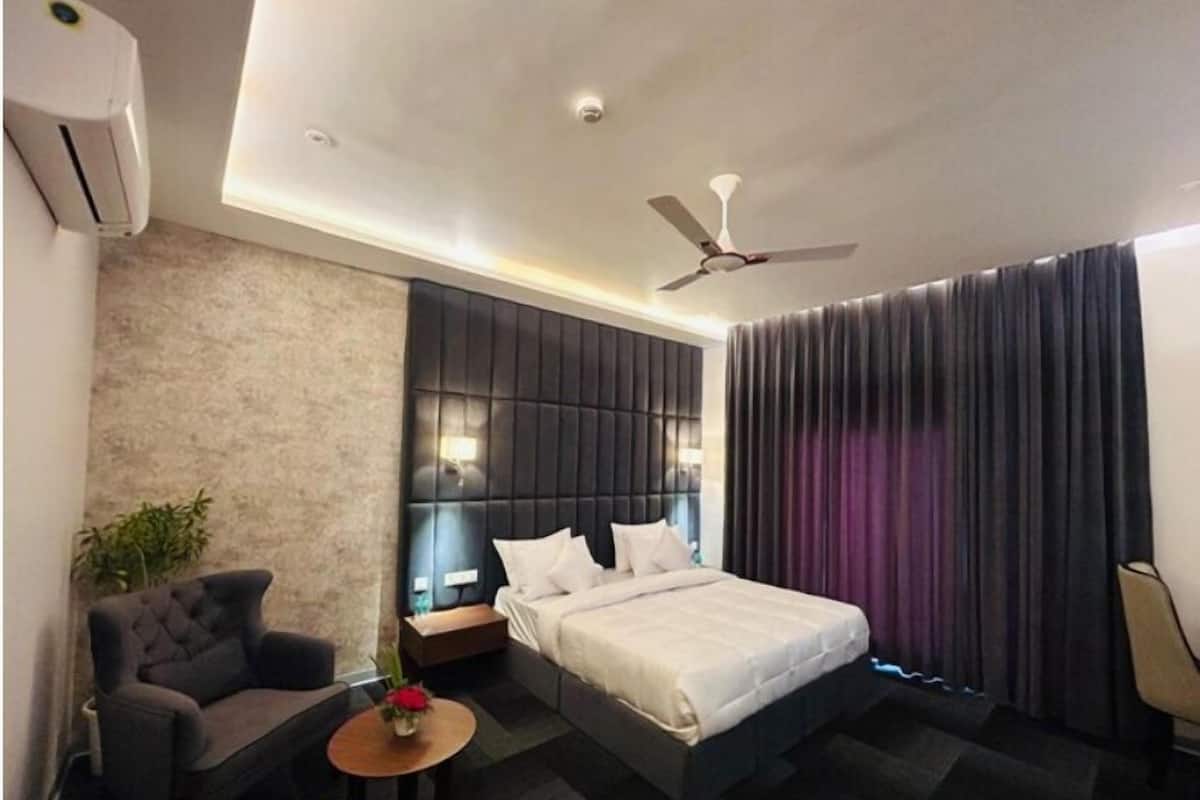 A modern hotel room with a bed, a seating area with a sofa and chairs, a small table with a vase, a ceiling fan, and a window with curtains.