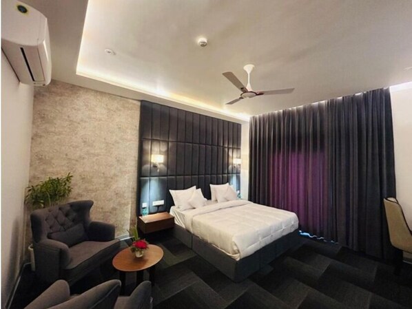 A modern hotel room with a bed, a seating area with a sofa and chairs, a small table with a vase, a ceiling fan, and a window with curtains. - Eminence the Corbett (Ramnagar)