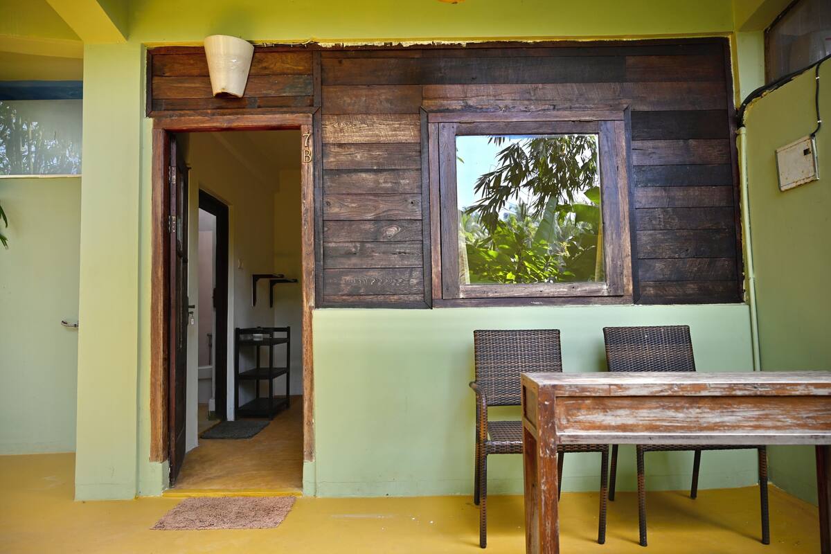A room with a wooden table and two wicker chairs, a window with a view of trees, and a door leading to another room.