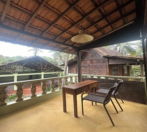 A covered outdoor area with a table and chairs, a thatched roof, and a view of a traditional building. - Sohum Spiritual Resort (Arambol)