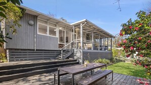 A covered outdoor area with a wooden deck, a bench, and a staircase leading to a building with large windows. - Nella (Lorne)