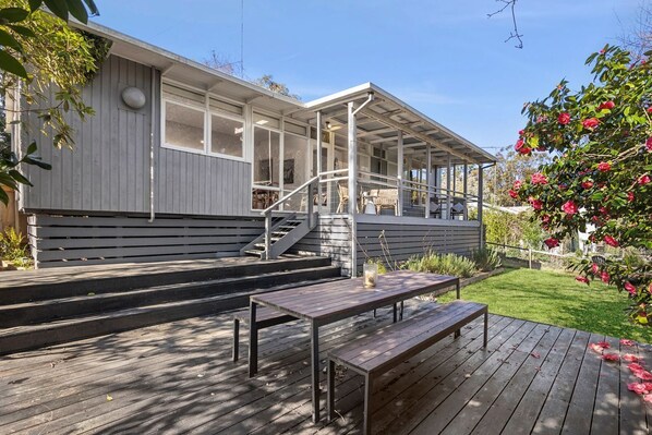 A covered outdoor area with a wooden deck, a bench, and a staircase leading to a building with large windows.