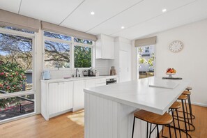 Een moderne keuken met witte kasten, een groot eiland met barkrukken en uitzicht op een tuin door de ramen.