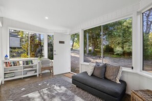 A cozy living room with a sofa, a bookshelf, and large windows offering a view of the outdoors.