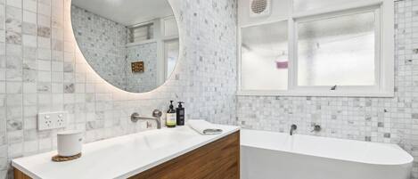 Un baño moderno con una bañera independiente, un espejo grande y un tocador blanco con un gabinete de madera.