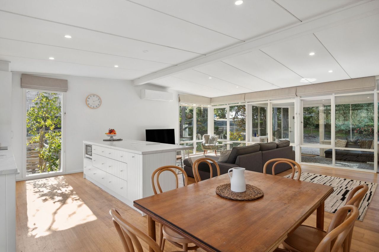 A modern kitchen with a dining area, large windows, and a view of greenery.