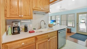 Una cocina con armarios de madera, una encimera blanca, cafetera, hervidor y pileta.