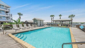 A pool area with a wooden deck, lounge chairs, a small building, and palm trees.