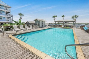 A pool area with a wooden deck, lounge chairs, a small building, and palm trees.