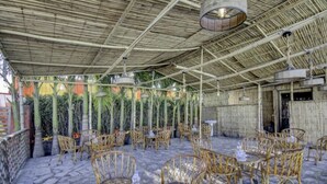 An outdoor patio area with wicker chairs and tables, bamboo ceiling, and a stone floor. - Parinay Vatika Banquet & Party Lawn (Haldwani)