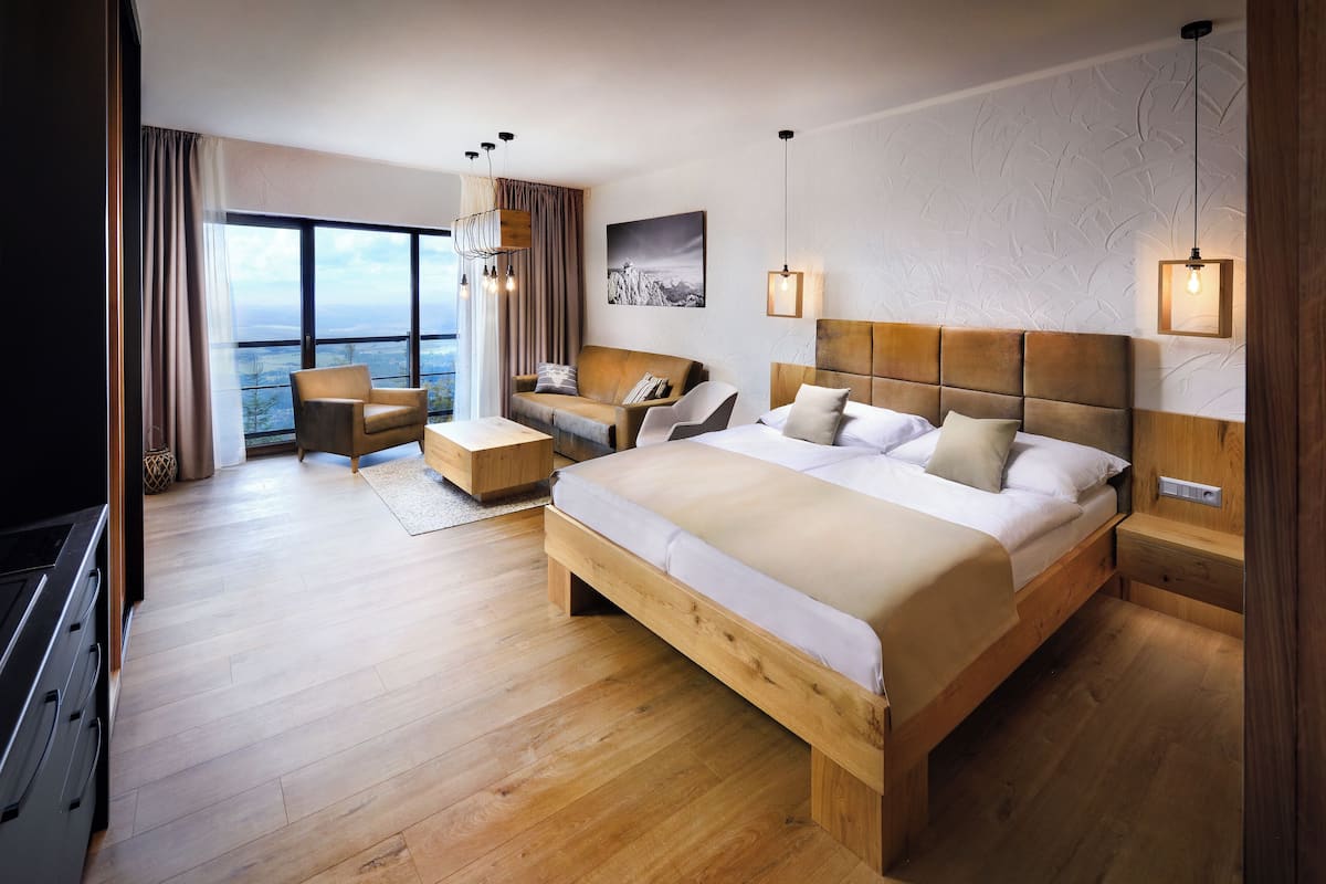 Habitación de hotel moderna con una cama grande, un sofá, una silla y un escritorio. Cuenta con un ventanal con vistas a las montañas.