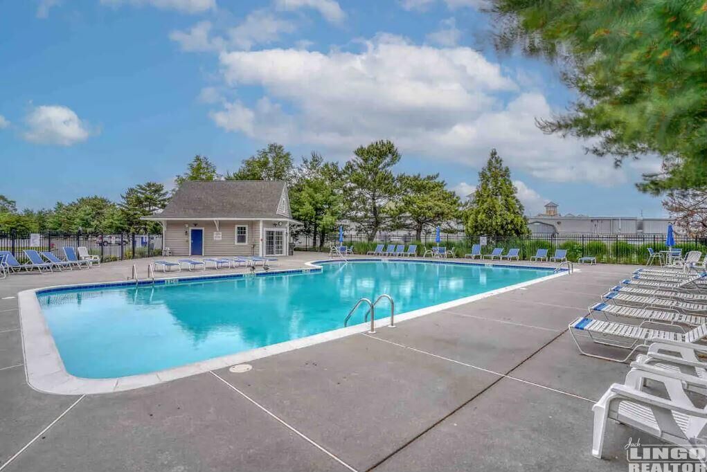 A rectangular swimming pool with a blue border, surrounded by lounge chairs and a small building in the background.
