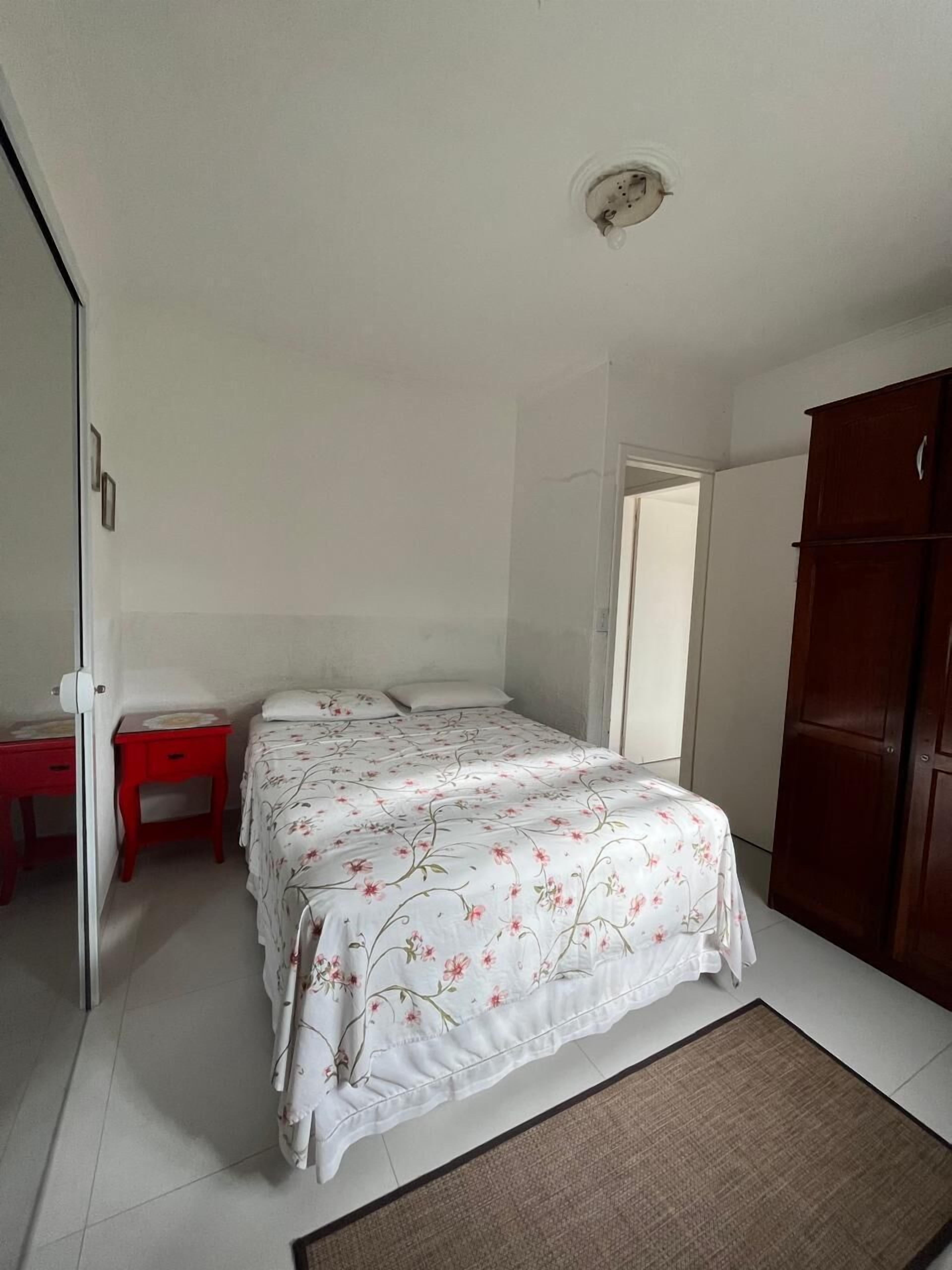 A bedroom with a bed, a red nightstand, a wooden wardrobe, and a door leading to another room.