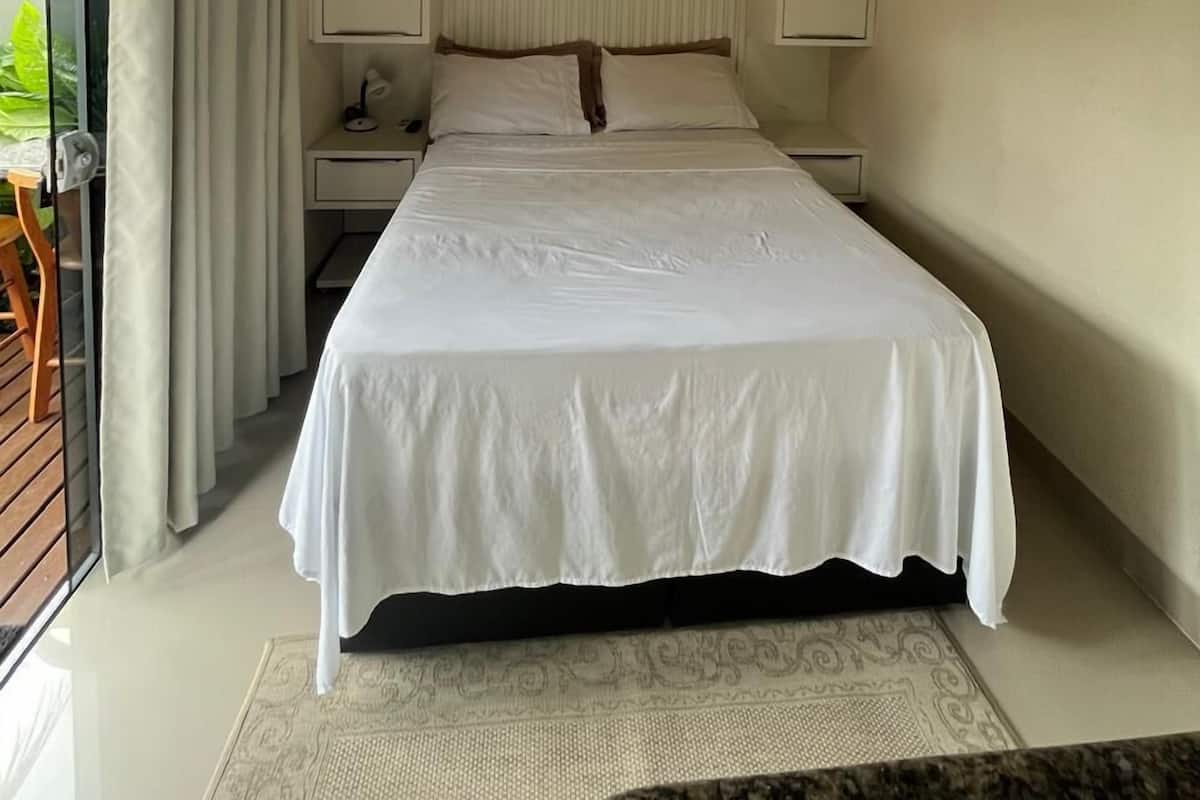 A bedroom with a wooden ceiling, a bed with white bedding, a nightstand, a desk, and a window with curtains.