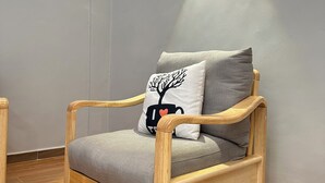 A wooden armchair with grey cushions and a decorative pillow against a grey wall, with a wooden floor and a small white rug. - Nsiimenta Stay (Kampala)