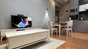 A modern living room with a flat-screen TV, a dining table with chairs, a kitchen area with cabinets, and a patterned rug on the wooden floor. - Nsiimenta Stay (Kampala)