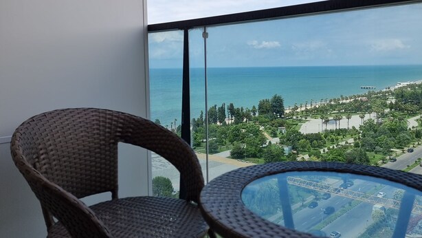 Ein Balkon mit Korbmöbeln, einem Glastisch und Blick auf das Meer und einen Park.