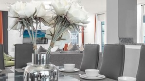A modern dining area with a glass table, white flowers in a vase, and grey upholstered chairs.