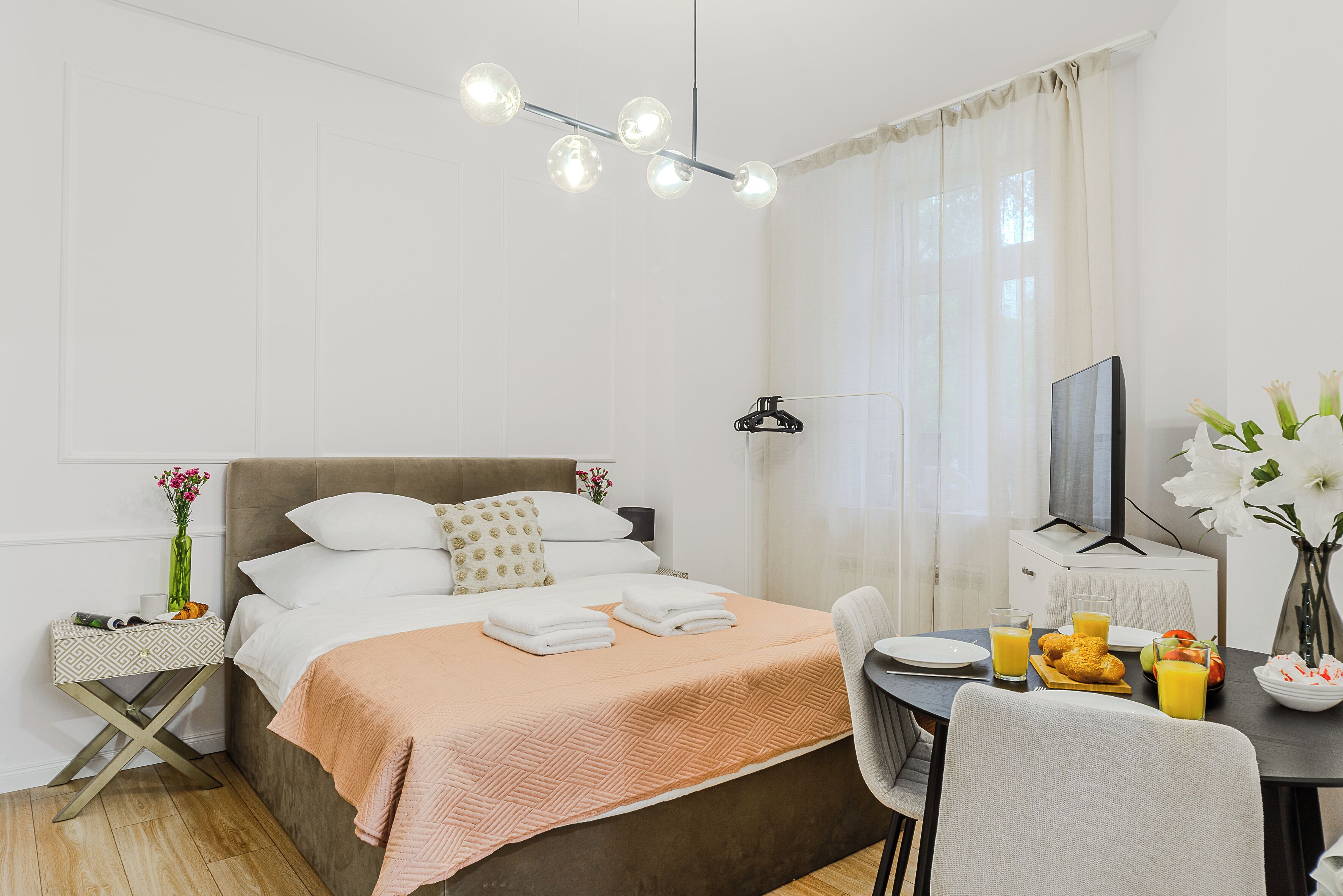 Habitación de hotel ordenada con cama, mesita de noche y zona de comedor con productos para el desayuno.