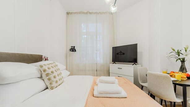 A hotel room with a bed, a TV, a desk with a chair, and a table with a vase of flowers. - Apartament Ogrodowa 26a (Warsaw)