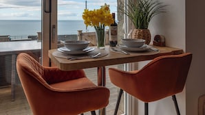 Una mesa de comedor con dos sillas, un jarrón con flores amarillas, una botella de vino y una copa en un balcón con vista al mar.