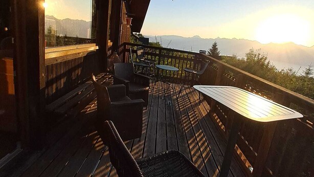 Eine Holzterrasse mit Außenmöbeln, einer Glaseinfriedung und Blick auf Berge.