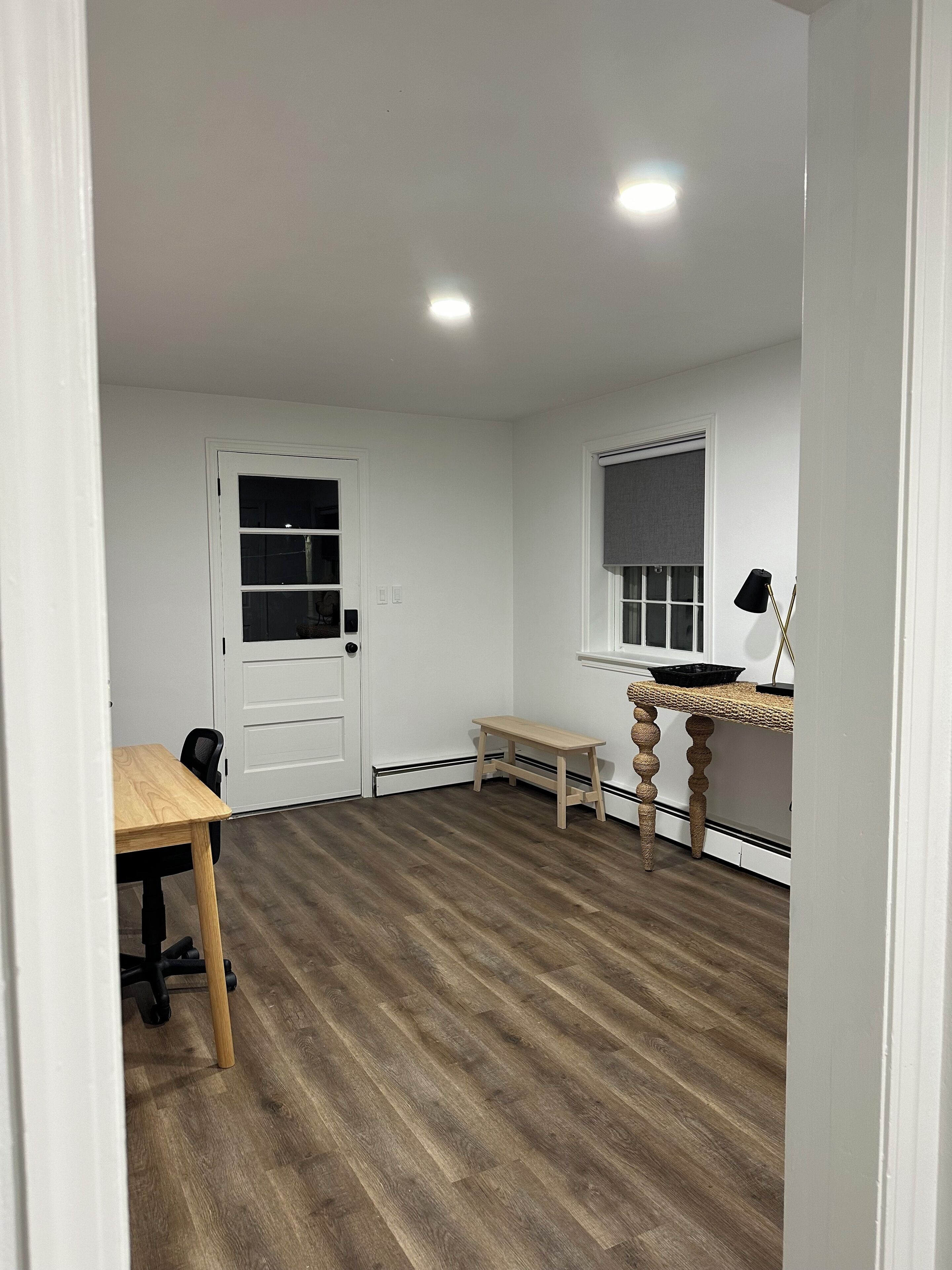 Una habitación con suelo de madera, una puerta blanca, una mesita y una ventana con persiana.