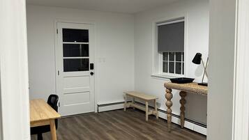 Una habitación con suelo de madera, una puerta blanca, una mesita y una ventana con persiana.