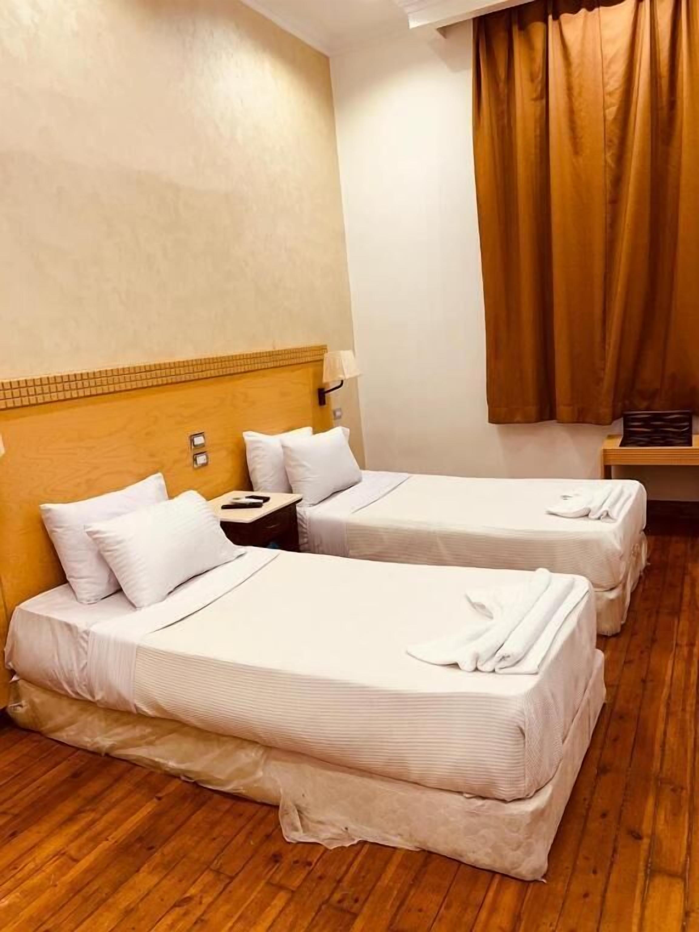 A hotel room with two beds, wooden flooring, a headboard with a light fixture, and a window with brown curtains.