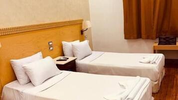 A hotel room with two beds, wooden flooring, a headboard with a light fixture, and a window with brown curtains.
