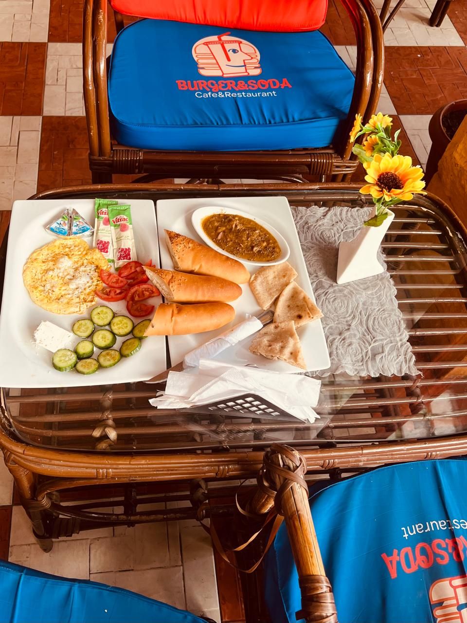 A dining table with a plate of food, including an omelette, bread, and vegetables, and a blue cushion with the Burger & Soda logo.