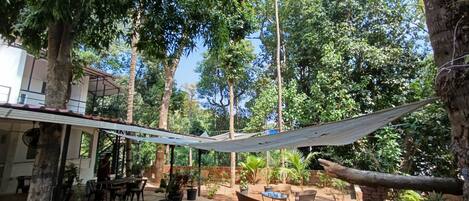 Uma área externa com mesas e cadeiras, cercada por plantas em vasos e árvores.