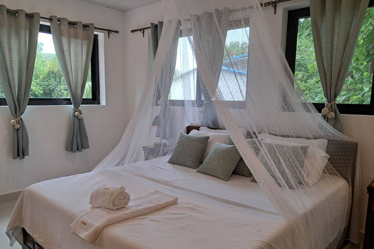 A neatly made bed with a white bedspread and pillows, a rolled towel on the side, and a mosquito net over the bed.