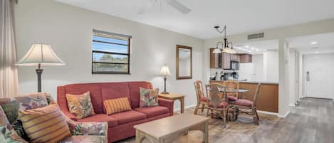 A living room with a red sofa, floral armchair, and a wooden coffee table. There is a ceiling fan, a dining area with a table and chairs, and a kitchen in the background.