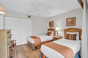Quarto de hotel com duas camas, um ventilador de teto, mesa de cabeceira de madeira com abajur e guarda-roupa com portas venezianas.