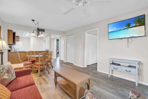 A spacious living area with a wooden table, a sofa, and a TV mounted on the wall displaying a beach scene.