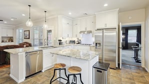 A modern kitchen with white cabinets, a stainless steel refrigerator, a built-in oven, and a large island with stools. - Vista Valley Vacay, 5-BD w Pool & Cinema (Lewisville)