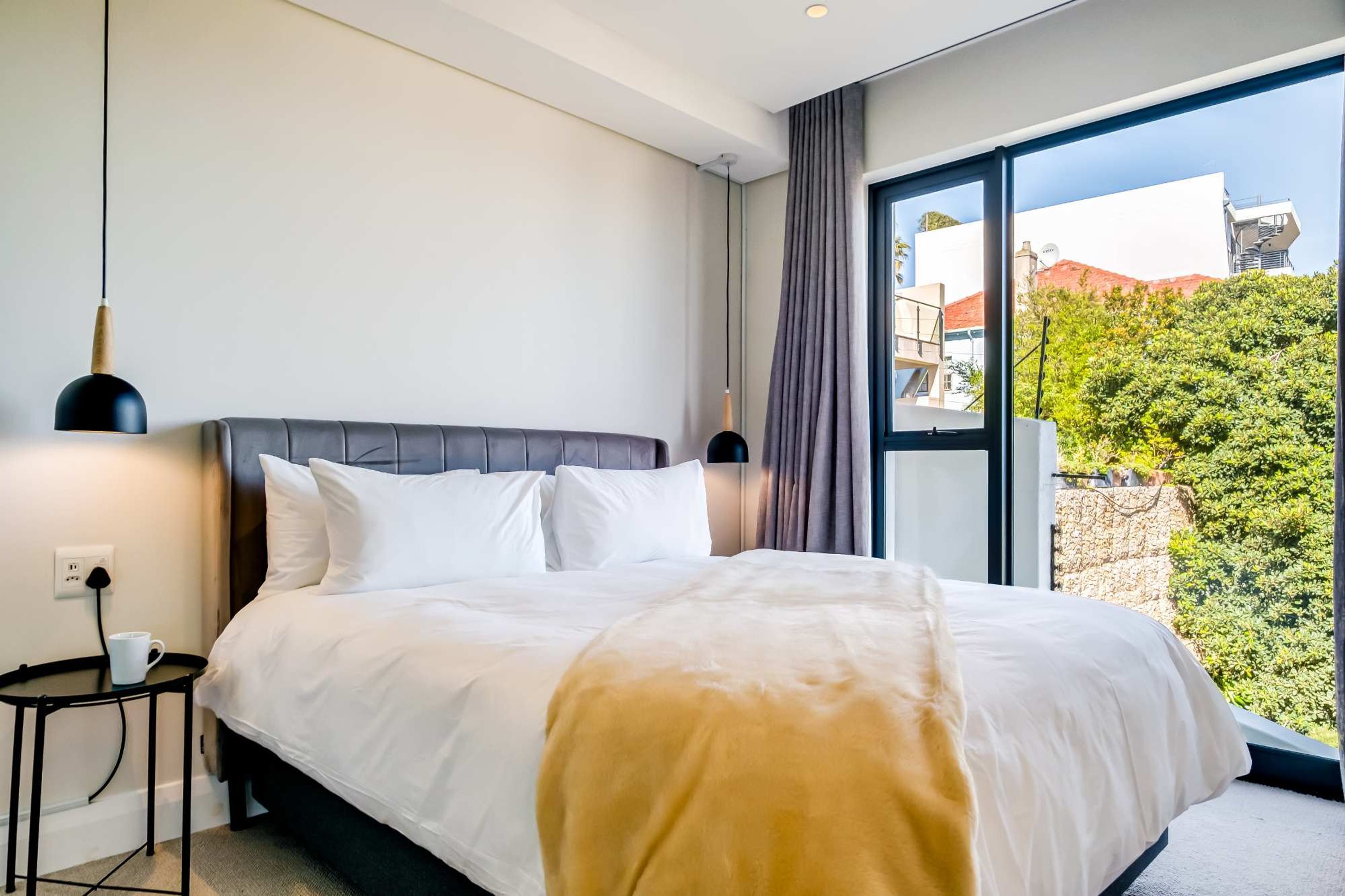 Ein modernes Hotelzimmer mit einem großen Bett, einem Nachttisch, einer Lampe und Blick auf Grünflächen und Gebäude.