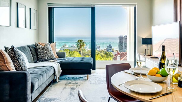 A modern living room with a sofa, a dining table set with plates and glasses, and a large window overlooking a cityscape and ocean. - Charming Coastal Escape Marula One (Cape Town)