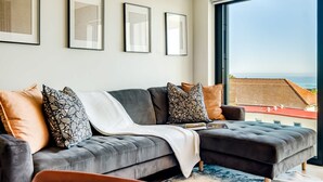 A modern living room with a grey sectional sofa, multiple cushions, and a large window offering a view of the city. - Charming Coastal Escape Marula One (Cape Town)