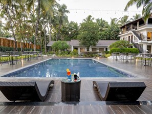 Zona de piscina con sillones de descanso, una barra y un edificio de dos pisos con balcones al fondo.