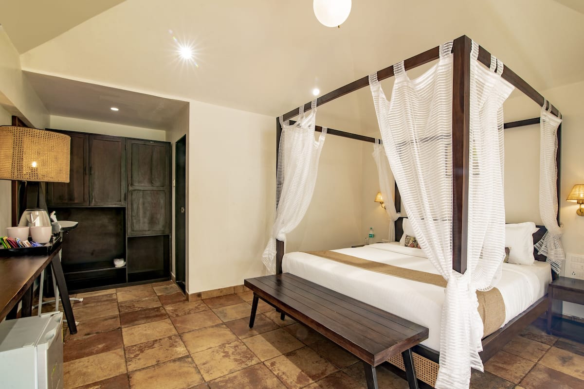 Una cama con dosel adornada con cortinas blancas, un banco de madera, una zona de cocina con armarios y una mesa de comedor con sillas.
