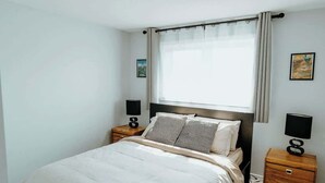 Ein Schlafzimmer mit einem Bett, zwei Nachttischen, einem Vorhangfenster und einem an der Wand montierten Fernseher.