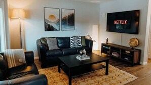 Ein Wohnzimmer mit einer schwarzen Ledersofa, einem Couchtisch, einem Fernseher, der 'NETFLIX' anzeigt, und einem Bücherregal.