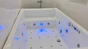 Un bain à remous rectangulaire blanc, doté de plusieurs jets et de lumières LED, installé devant un mur sobre agrémenté d’une petite fenêtre.