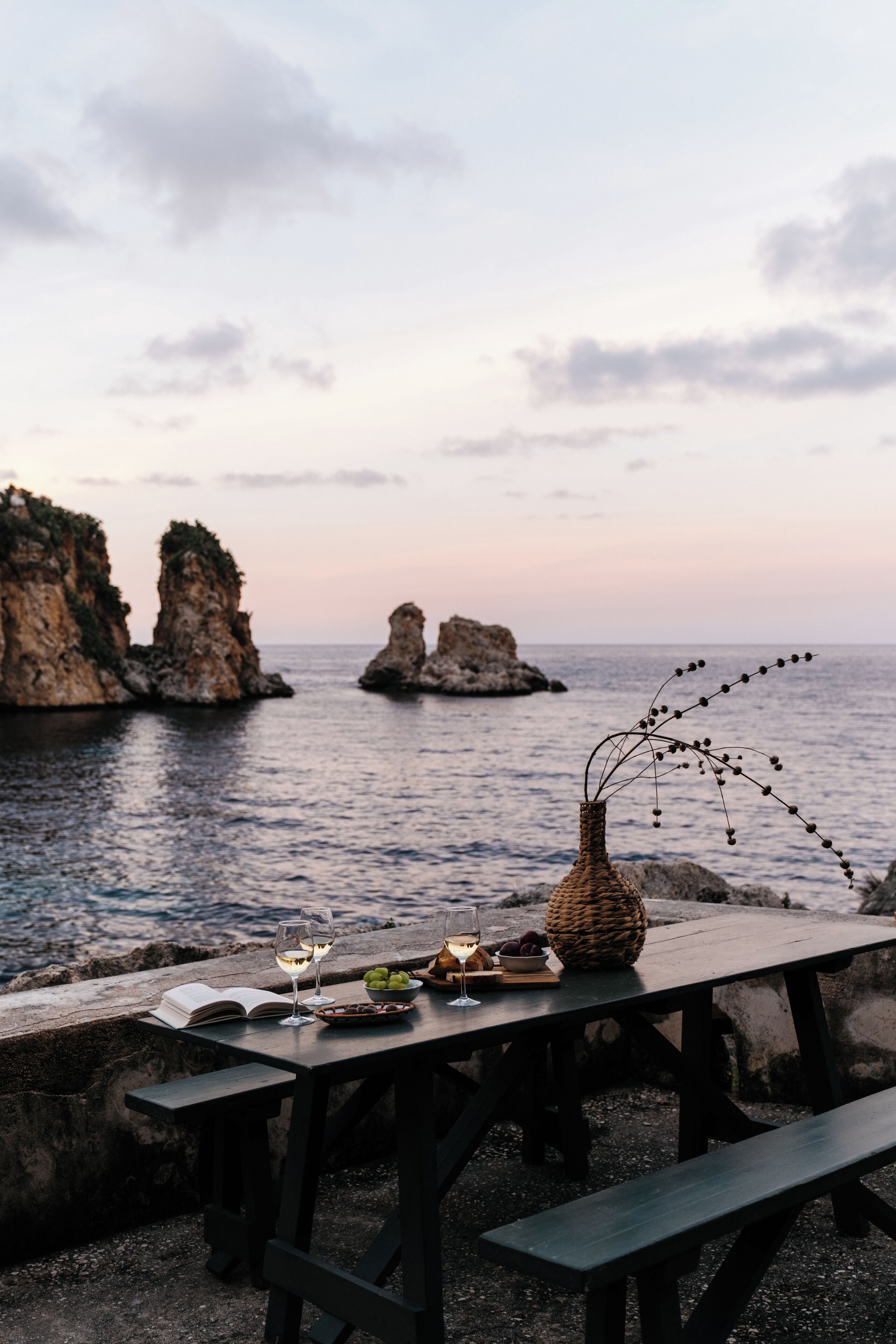 A wooden table with a vase, wine glasses, and fruits, overlooking a rocky coastline with clear skies.