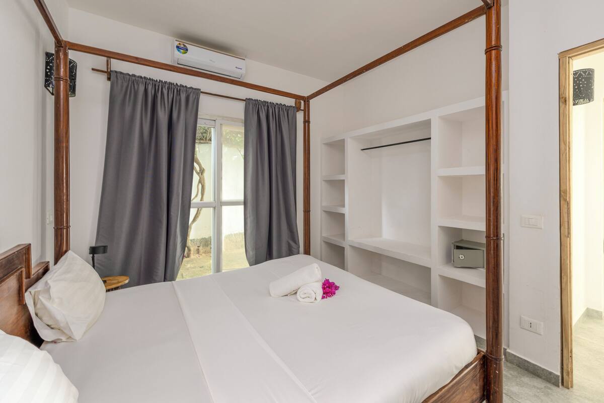 A hotel room with a large bed, grey curtains, a wardrobe, and an air conditioning unit.