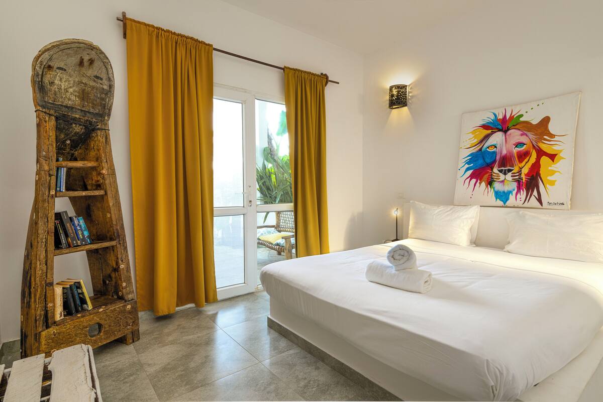 A hotel room with a bed, a wooden ladder bookshelf, yellow curtains, and a colorful lion painting.
